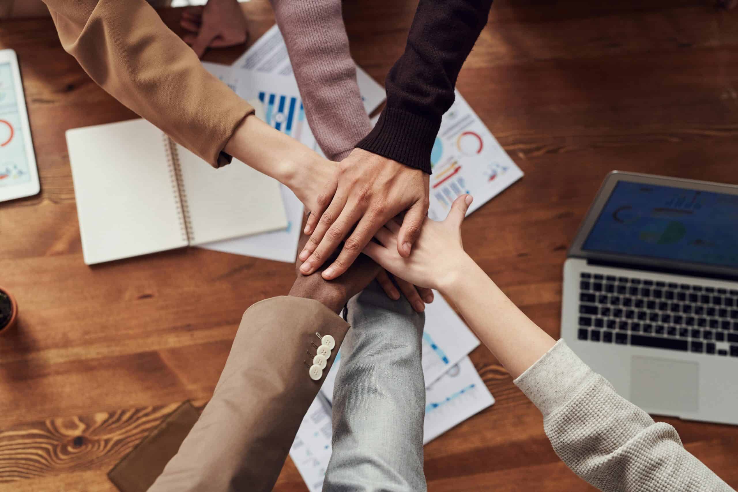 Arms of a diverse group of people in a huddle with hands stacked one on top of the other over a desk in a professional setting.