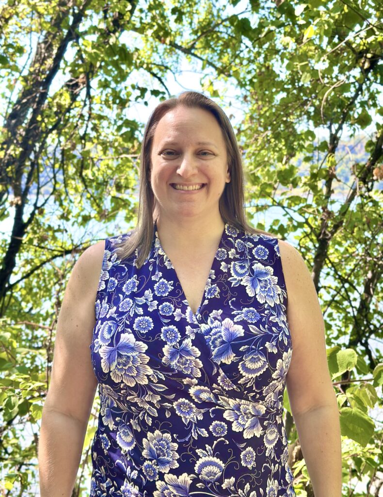 Mindfulness coach and meditation teacher, Robyn Gold standing in front of trees and a body of water, smiling with her arms relaxed at her sides.